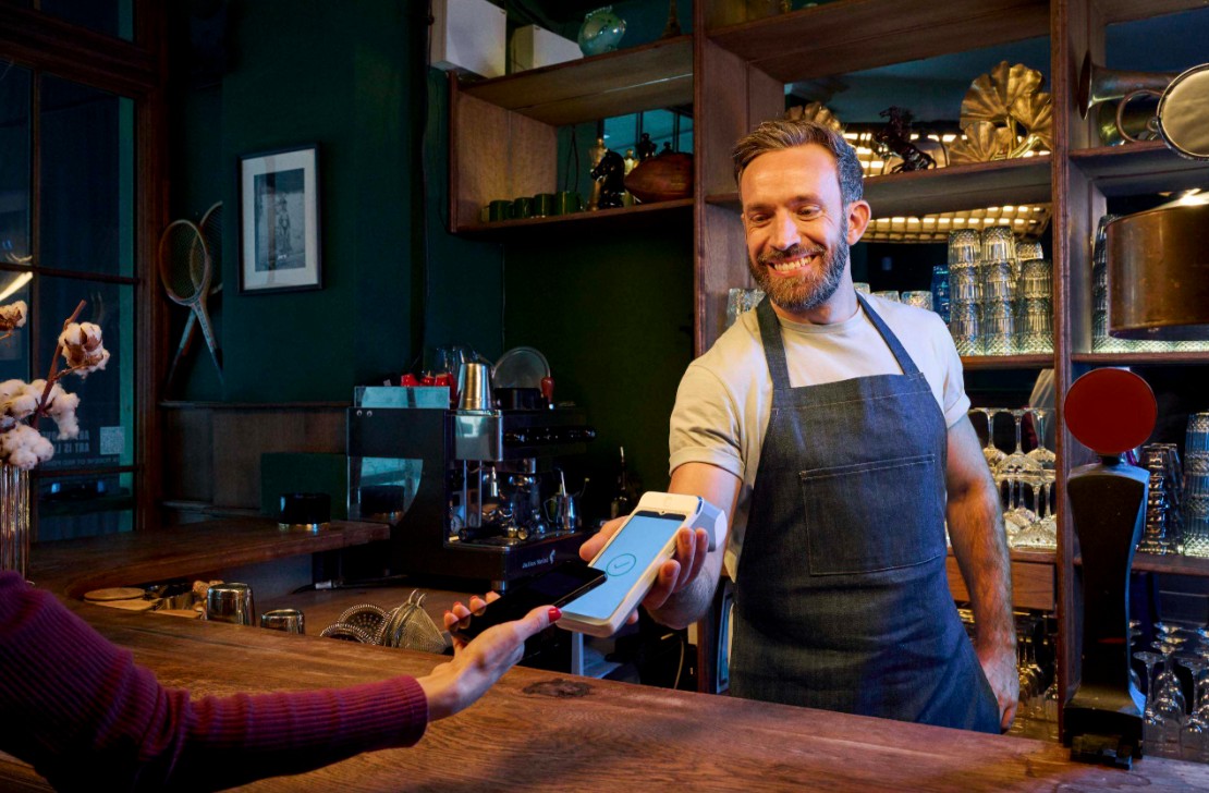 Barista in an apron hands a payment terminal to a customer at a wooden bar.